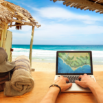 Man’s hand on a laptop on a table near a beach, with a backpack and a wooden house in the background, overlooking the sea.