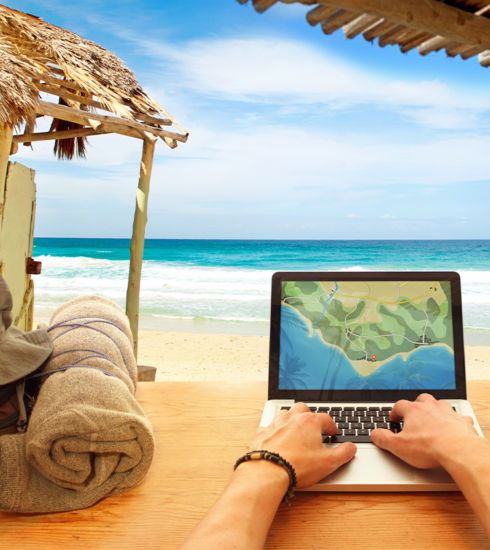 Man’s hand on a laptop on a table near a beach, with a backpack and a wooden house in the background, overlooking the sea.