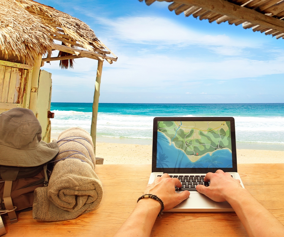 Man’s hand on a laptop on a table near a beach, with a backpack and a wooden house in the background, overlooking the sea.